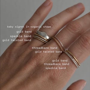 May include: A hand displays a collection of rings in various styles. The rings include a baby signet in an organic shape, a gold band, a sparkle band, and a gold twisted band. Other rings are threadbare and gold.
