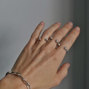May include: A hand displays several silver rings with spherical accents. The rings are worn on multiple fingers, showcasing a minimalist design. A silver chain bracelet is also visible on the wrist, complementing the rings. The background is a plain gray.