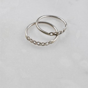 May include: Two silver rings with a chain link design. The rings are thin and delicate, with a simple yet elegant style. The rings are displayed on a white marble surface.
