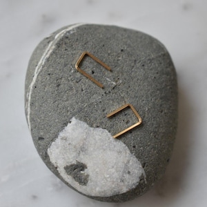 May include: A pair of gold minimalist earrings in the shape of a rectangle. The earrings are displayed on a gray and white rock.