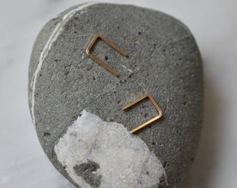 Op de afbeelding: Een paar gouden minimalistische oorbellen in de vorm van een rechthoek. De oorbellen worden gepresenteerd op een grijze en witte steen.