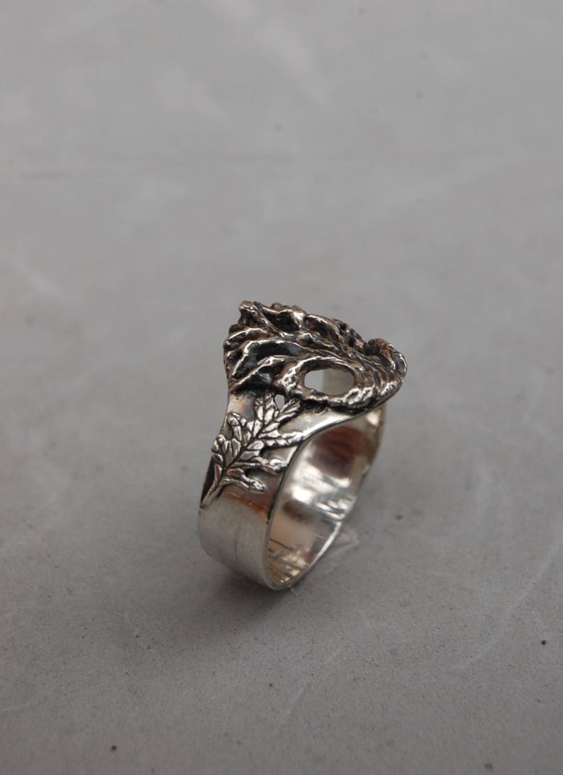 May include: A silver ring with a detailed design of a tree with leaves. The ring is made of metal and has a smooth finish.