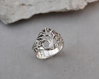 May include: A silver ring with a tree design. The tree is intricately detailed and has a cutout in the center. The ring is a wide band and has a polished finish.