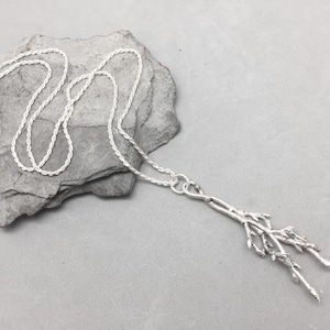 Peut inclure: Un collier argenté avec une chaîne torsadée et un pendentif en forme de branches stylisées. Le collier est posé sur une pierre grise sur fond gris. Le pendentif présente des détails fins.