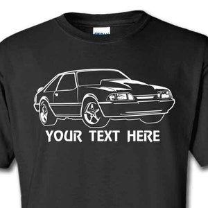 May include: Black t-shirt featuring a white outline of a Ford Mustang car. The text "YOUR TEXT HERE" is printed below the car.