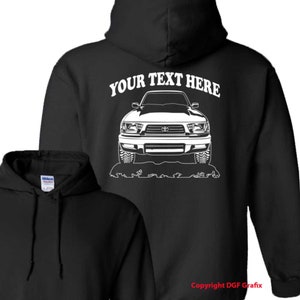 May include: Black hooded sweatshirt with a white outline of a Toyota Tacoma truck on the back. The text "YOUR TEXT HERE" is above the truck. The sweatshirt is a blank canvas for personalization.