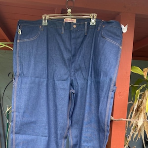 May include: Dark blue denim work pants with orange stitching. The pants have a classic five-pocket design, a straight leg, and a metal button closure. The brand name "Big Mac" is visible on the waistband.