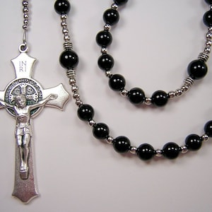 May include: A silver rosary with black beads and a silver crucifix featuring the image of Saint Francis of Assisi. The crucifix has the inscription "IN RI" on the top.