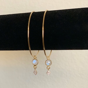 Puede incluir: Dos pulseras de aro de oro con un colgante de piedra lunar y un colgante en forma de diamante.