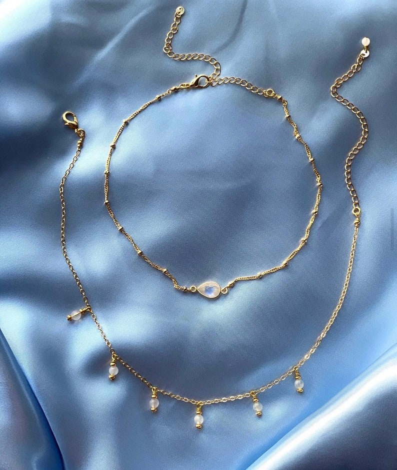 K&ouml;nnte beinhalten: Zwei goldene Halsketten mit zarten Ketten. Die obere Halskette hat einen tropfenf&ouml;rmigen Mondstein-Anh&auml;nger. Die untere Halskette hat eine Reihe kleiner wei&szlig;er Perlen, die daran h&auml;ngen.