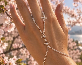 May include: A silver hand chain featuring three delicate flower-shaped links, each centered with a small clear stone. The jewelry connects a ring loop to a wrist chain, constructed from fine metallic links that drape across the back of the hand.
