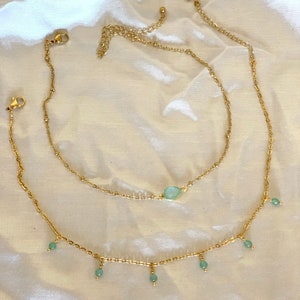 K&ouml;nnte beinhalten: Zwei goldene Kettenhalsketten mit einem hellblauen Edelstein-Anh&auml;nger an der oberen Kette und hellblauen Perlen, die von der unteren Kette h&auml;ngen.