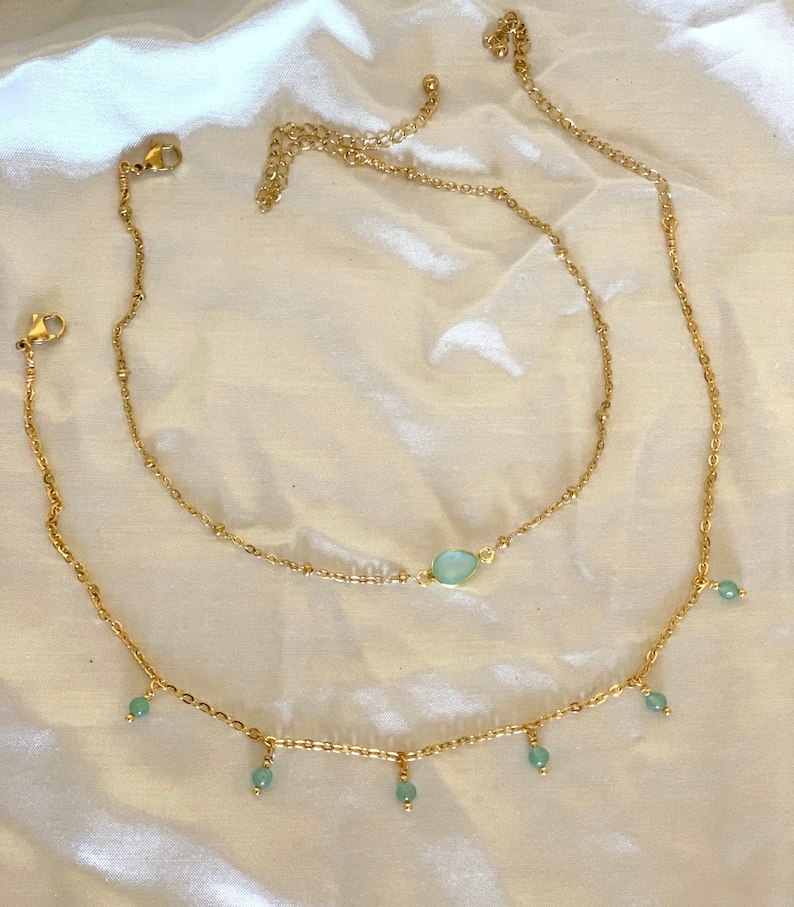 K&ouml;nnte beinhalten: Zwei goldene Kettenhalsketten mit einem hellblauen Edelstein-Anh&auml;nger an der oberen Kette und hellblauen Perlen, die von der unteren Kette h&auml;ngen.