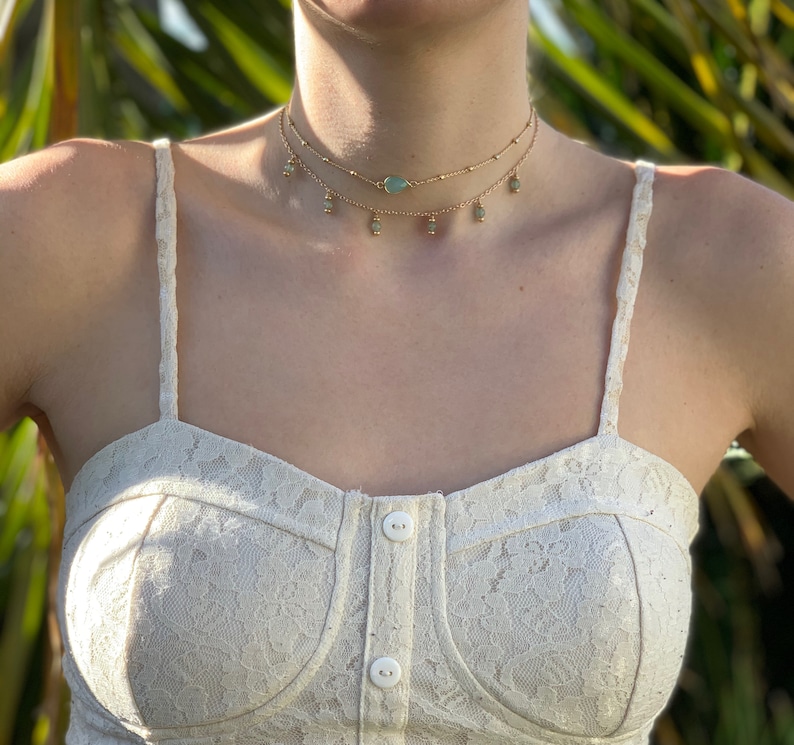 K&ouml;nnte beinhalten: Eine goldene mehrreihige Choker-Kette mit einem tropfenf&ouml;rmigen Anh&auml;nger aus gr&uuml;nem Stein und kleinen runden gr&uuml;nen Steinen, die an der Kette baumeln.
