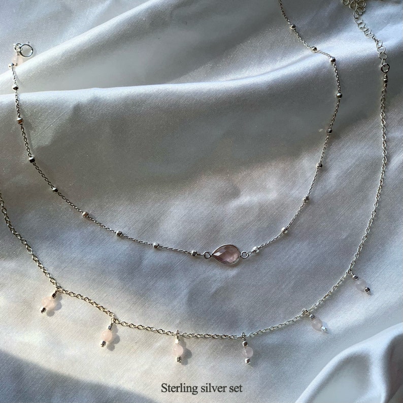 K&ouml;nnte beinhalten: Ein zartes Silber-Halsketten-Set mit einem rosa Edelstein-Anh&auml;nger und kleineren rosa Edelstein-Akzenten. Die Halskette ist mit einer k&uuml;rzeren Kette und einer l&auml;ngeren Kette mit baumelnden Anh&auml;ngern geschichtet. Sterling Silber Set.
