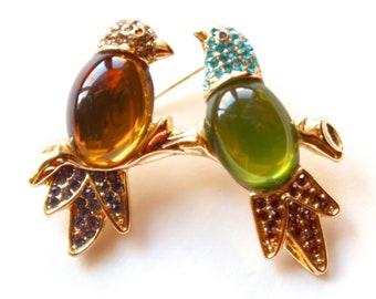 May include: A gold-tone brooch featuring two birds perched on a branch. The birds have jeweled eyes and are adorned with colorful rhinestones. One bird has an amber-colored body and the other has a green body.