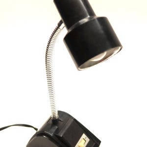 Puede incluir: Lámpara de escritorio negra con cuello flexible de color plateado. La lámpara tiene una base negra y una pantalla negra. El interruptor de encendido/apagado es visible en la base. La lámpara está diseñada para colocarse en un escritorio o mesa.