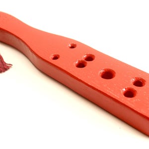May include: A red wooden paddle with multiple circular holes and a knotted red string attached to the handle. The paddle has a smooth, glossy finish and is set against a white background.