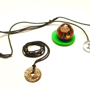May include: A collection of handcrafted necklaces and a decorative doll. One necklace features a coin pendant, another a silver disc with the Roman numeral "II", and the third a doll on a green base. The necklaces have black cords.