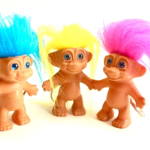 May include: Three vintage Troll dolls with colorful, spiky hair. The dolls have tan skin, blue eyes, and are smiling. One has blue hair, one has yellow hair, and the other has pink hair. They are standing on a white surface.