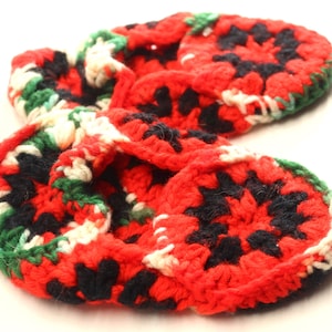 May include: A handmade crochet item featuring a repeating pattern of red, black, white, and green. The item appears to be a scarf or a similar accessory, with a soft, textured appearance. The design is a series of connected circular shapes.