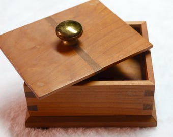 May include: A wooden box with a hinged lid and a brass knob. The box is made of light brown wood with a darker wood inlay. The box is closed.