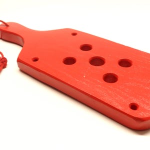 May include: A bright red wooden paddle with a handle and several circular holes. A red string is attached to the handle. The paddle is on a white background.