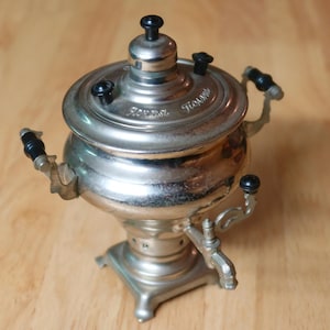 May include: A silver samovar with black handles and a spout. The samovar has a round body and a domed lid with a knob on top. The samovar is sitting on a wooden surface.