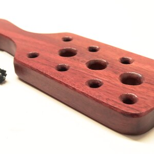 May include: A wooden paddle with a dark red finish and a rope handle. The paddle has multiple round holes of varying sizes.