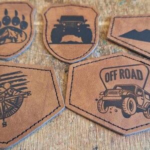 May include: Five brown leather patches with different designs. The designs include a bear paw print, a jeep, mountains, an American flag with a compass, and a jeep with the words "Off Road".