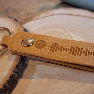 May include: A tan leather keychain with a gold metal key ring. The keychain has a small metal rivet and a laser-engraved Spotify logo and a sound wave pattern.