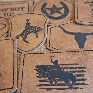 May include: A collection of nine brown leather patches with various designs, including a cowboy riding a horse, a wolf howling at the moon, a skull, a horseshoe with a star, a boot with flowers, and the text "RANCH LIFE" and "COWBOY UP".