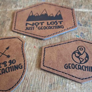 May include: Three brown leather patches with black stitching and text. The first patch says "Not Lost Just Geocaching" with a mountain range graphic. The second patch says "Let's Go Geocaching" with two crossed arrows. The third patch says "Geocaching" with a globe and pin graphic.