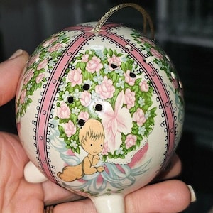 Peut inclure: Une boule de Noël en céramique blanche avec un motif floral rose et vert. La boule est munie d'une ficelle dorée pour l'accrocher et de trois petits pieds. Un petit chérubin est peint sur la boule.