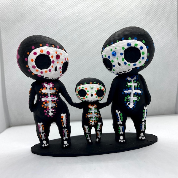 Può includere: Una famiglia di tre figurine di scheletro dipinte in bianco e nero. Le figurine sono in piedi su una base nera. Le figurine hanno punti colorati sui loro corpi e volti.