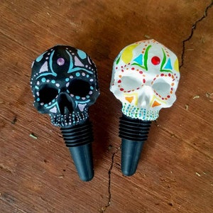 May include: Two decorative skull-shaped bottle stoppers. One is black with blue and purple accents, the other is white with colorful designs. Both have black rubber stoppers and are set against a wooden background.