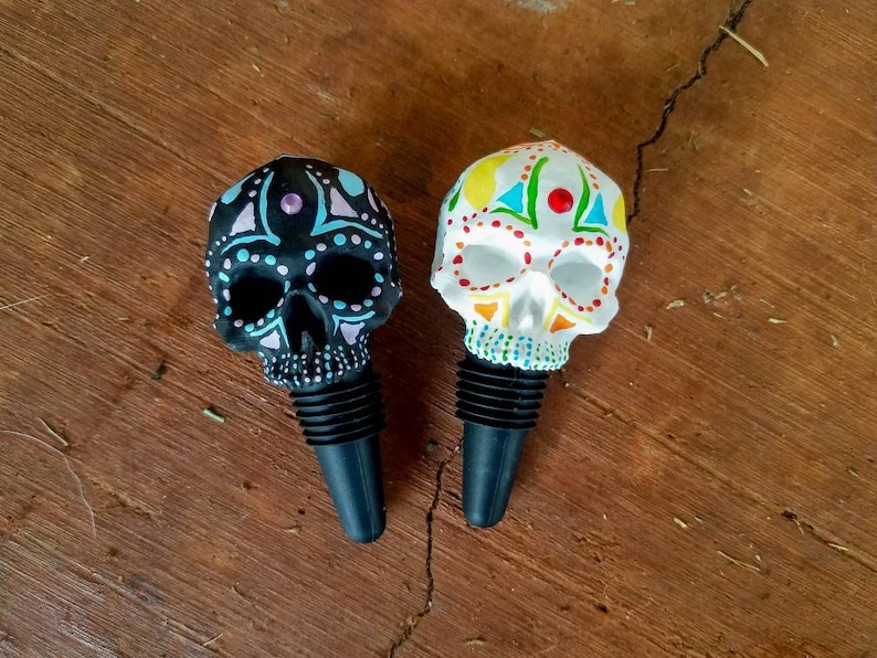 May include: Two decorative skull-shaped bottle stoppers. One is black with blue and purple accents, the other is white with colorful designs. Both have black rubber stoppers and are set against a wooden background.