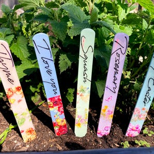 Puede incluir: Cinco marcadores de plantas de madera coloridos con etiquetas escritas a mano: Tomillo, Te amo, Espinacas, Romero y Feliz Pascua. Los marcadores están decorados con motivos florales.