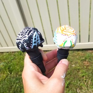 May include: Two decorative skull-shaped bottle stoppers. One is black with white and light blue designs, the other is white with colorful floral patterns. Both have black rubber bases.