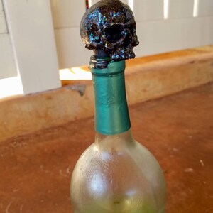 May include: A black skull bottle stopper with iridescent glitter sits atop a green-necked wine bottle. The clear glass bottle has condensation. The stopper is a decorative item for wine bottles.