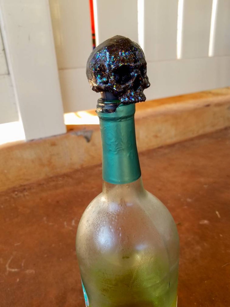 May include: A black skull bottle stopper with iridescent glitter sits atop a green-necked wine bottle. The clear glass bottle has condensation. The stopper is a decorative item for wine bottles.