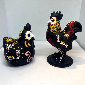 May include: Two black ceramic figurines: a hen and a rooster. Both are decorated with colorful Day of the Dead-style designs. The hen is squatting, while the rooster stands on a round base. The rooster has a tall comb.