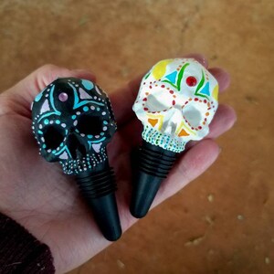 May include: Two decorative skull-shaped bottle stoppers. One is black with blue and pink accents, the other is white with colorful geometric designs. Both have black rubber stoppers, perfect for wine bottles.