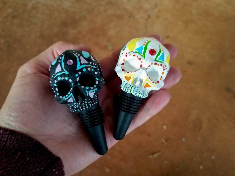May include: Two decorative skull-shaped bottle stoppers. One is black with blue and pink accents, the other is white with colorful geometric designs. Both have black rubber stoppers, perfect for wine bottles.