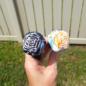 May include: Two decorative bottle stoppers. One is black with a white rose design and the other is white with an orange rose design. Both are hand-painted with colorful accents.