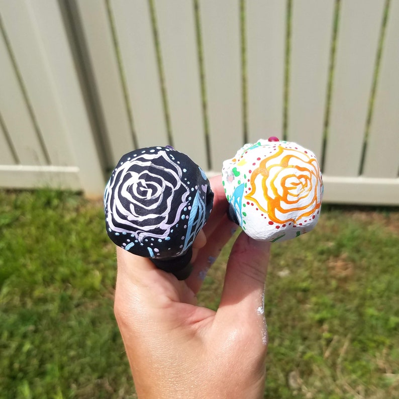 May include: Two decorative bottle stoppers. One is black with a white rose design and the other is white with an orange rose design. Both are hand-painted with colorful accents.