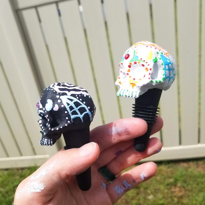 May include: Two decorative skull bottle stoppers. One is black with white and light blue accents, the other is white with colorful floral designs. Both have black rubber stoppers.