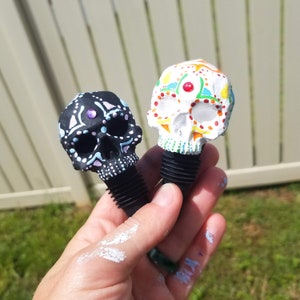May include: Two decorative skull-shaped bottle stoppers. One is black with white and light blue accents, the other is white with colorful floral designs. Both have black rubber stoppers. The stoppers are held in a hand.