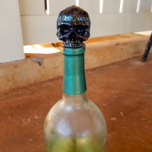 May include: A clear glass wine bottle with a teal green neck and a glittery black skull bottle stopper. The skull is covered in iridescent glitter, and the bottle contains a yellow liquid.