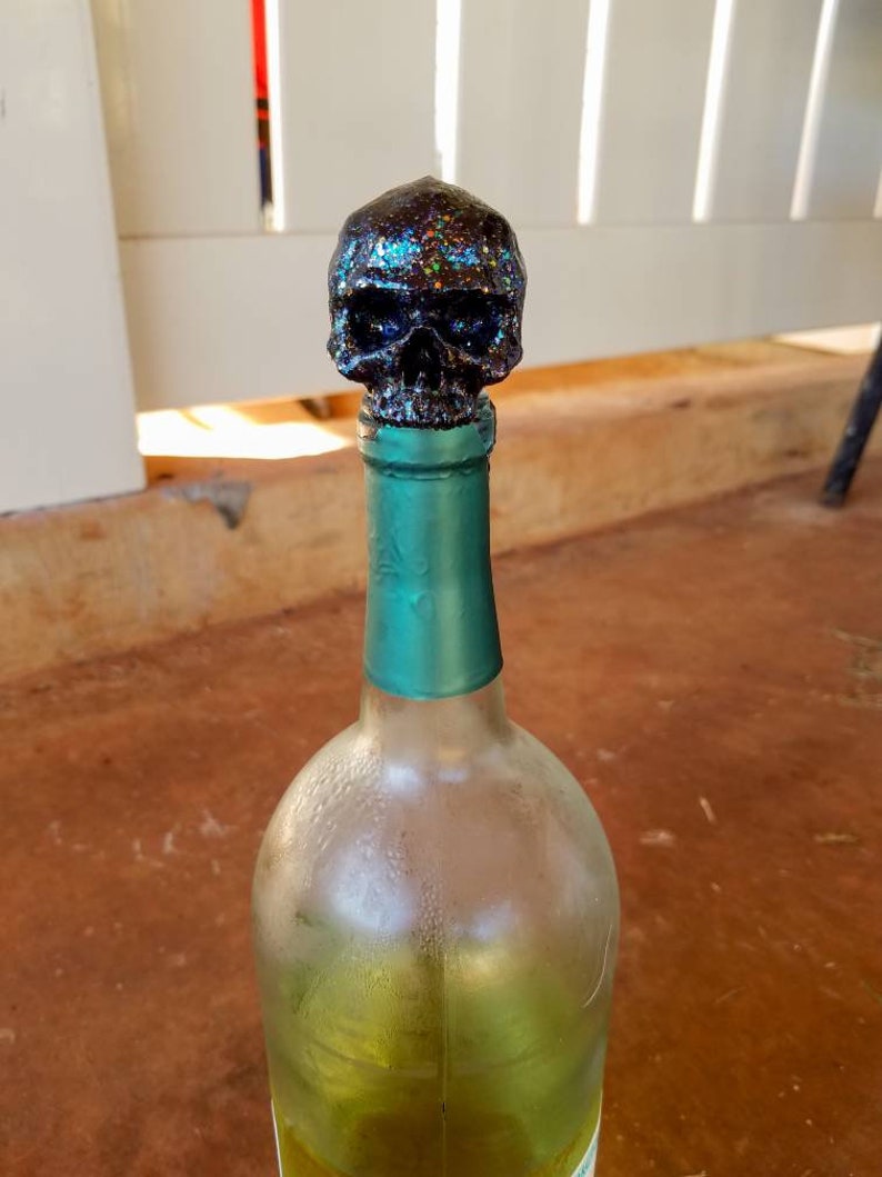 May include: A clear glass wine bottle with a teal green neck and a glittery black skull bottle stopper. The skull is covered in iridescent glitter, and the bottle contains a yellow liquid.